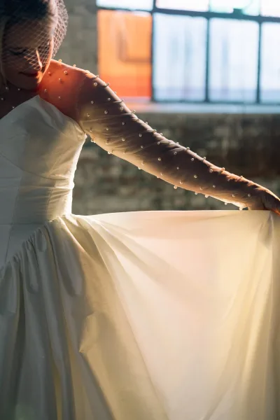 Bridal portrait of a bride in a satin wedding dress with birdcage veil, holding her skirt by sunlit industrial windows and concrete wall