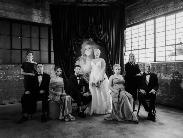 Wedding party portrait with bride centered holding a simple bridal bouquet, surrounded by bridesmaids and groomsmen in tuxedos by brick wall and windows