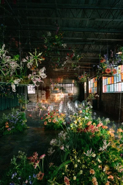 Ceremony aisle decor with a wildflower meadow runner, hanging floral installations, and clear acrylic chairs in a brick warehouse