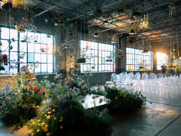 Ceremony setup with hanging floral installations and wildflower arrangements lining an aisle in a sunlit industrial loft with brick walls and factory windows