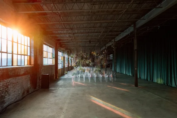 Indoor ceremony setup with clear acrylic chairs lining an aisle beneath hanging floral installation, candlelight and string lights in a brick warehouse venue