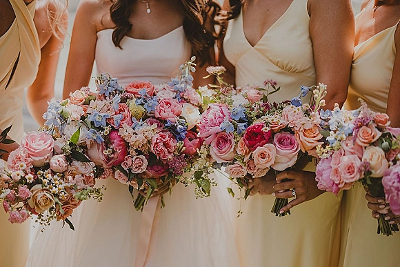 Bridesmaid bouquets in pastel mixed flowers with roses, peonies and delphinium, tied with ribbons, held in warm sunlight outdoors