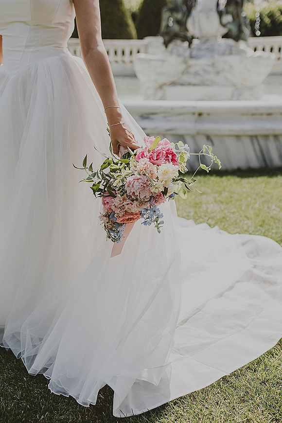 Bridal bouquet of pink peonies with greenery and ribbon held against a tulle wedding dress, with a stone garden fountain behind