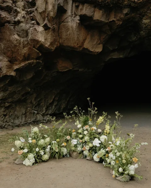 Wedding ceremony florals with grounded ceremony flowers in white and yellow blooms and greenery arranged on sand among rocks in a cave setting