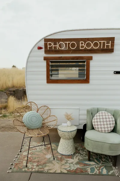 Wedding photo booth vintage trailer with neon sign and boho lounge seating, rattan and tufted chairs on a patterned rug in a grassy field