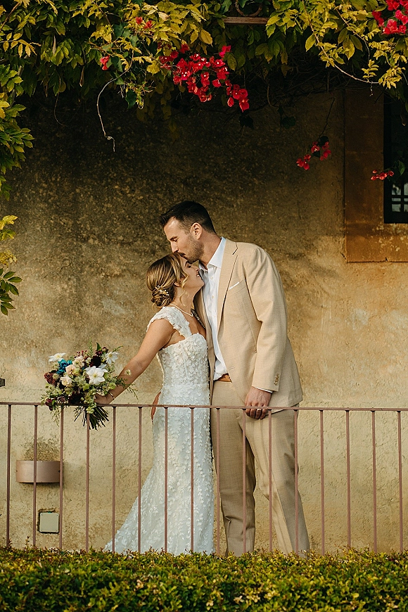 Couple portrait of groom kissing bride’s forehead as she holds a wedding bouquet on a balcony beside a stone wall with vines and red flowers