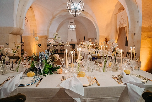 Reception tablescape with wedding table decor, taper candles and floral bud vases on white linen beneath an arched stone ceiling and lanterns