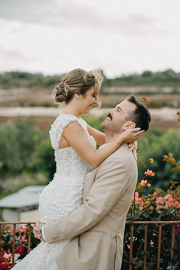Couple portrait of bride and groom embrace as he lifts her, lace gown and pearl earrings framed by terrace railing and cloudy hills backdrop