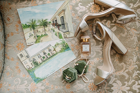 Bridal details flatlay featuring wedding shoes flatlay with silver strappy heels, pearl necklace, perfume, rings, and illustrated cards on patterned upholstery