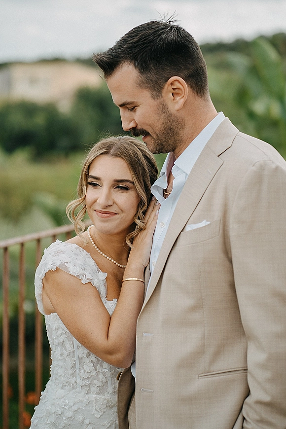 Couple portrait of bride and groom embrace, bride leaning on groom in lace dress and pearls by a wooden railing with hills and cloudy sky