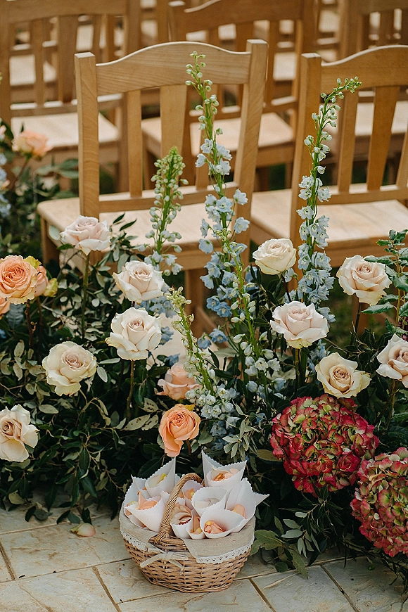 Ceremony floral decor with aisle floral arrangement of roses, blue delphinium and eucalyptus beside wood chairs on a stone patio