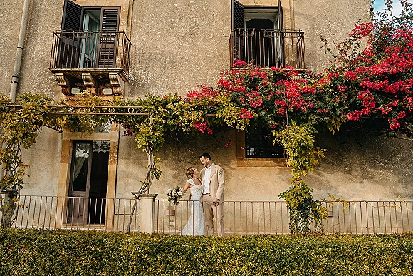 Couple portrait of bride and groom on balcony, bride holding bouquet beneath pink flowering vines against a stucco villa facade