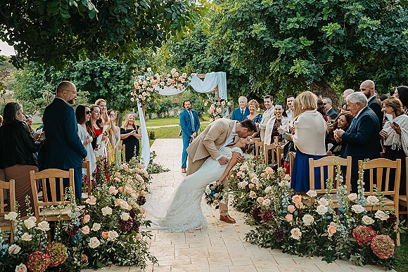 Wedding kiss moment as groom dips bride for a ceremony recessional kiss beneath a floral arch, guests cheering on a garden lawn aisle