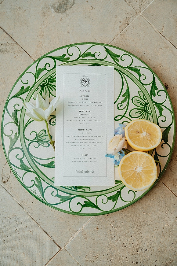 Wedding menu card styled on a green patterned dinner plate with citrus slices and a floral sprig on a stone tile floor