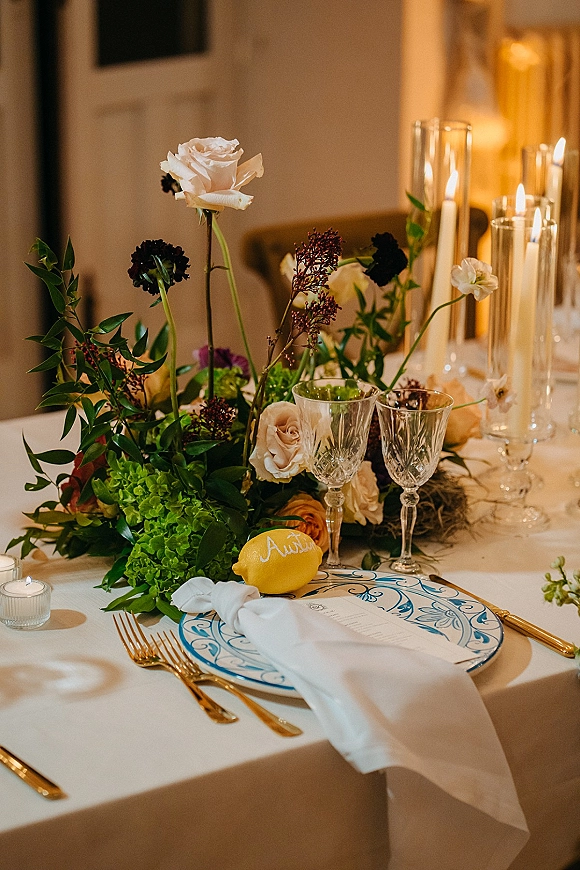 Wedding tablescape reception table setting with roses, hydrangea and greenery, taper candles, crystal goblets, gold flatware and lemon place card in warm interior lighting