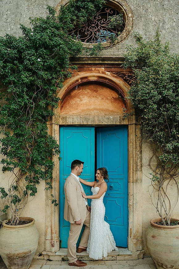Couple portrait of bride touching groom in a strapless lace gown and tan suit beside blue double doors framed by ivy and stone wall