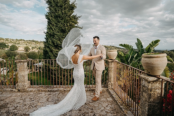 Couple portrait of bride and groom holding hands as her veil lifts in the wind on a stone terrace with tropical foliage and hills