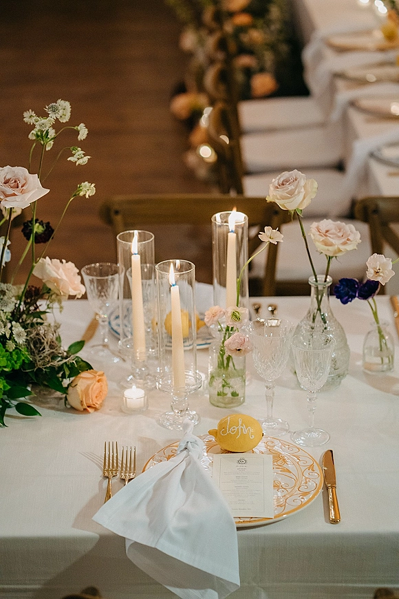 Reception tablescape wedding table setting with blush rose bud vases, gold flatware, crystal glassware, and taper candles on white linen by wood wall