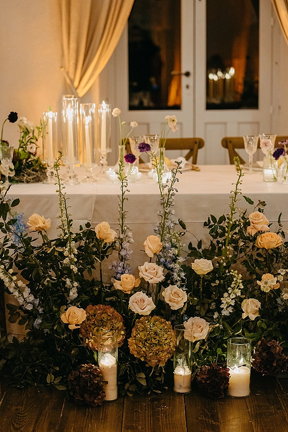 Reception sweetheart table with sweetheart table flowers in a low rose, hydrangea, and greenery meadow, surrounded by pillar and taper candles indoors