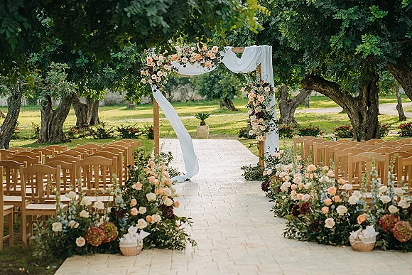 Ceremony setup for an outdoor wedding ceremony with wooden arch draped in white fabric and floral aisle arrangements on a stone path in sunny garden lawn