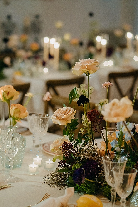 Reception tablescape with a wedding table centerpiece of roses, mixed florals and moss, surrounded by taper and votive candles on a white cloth