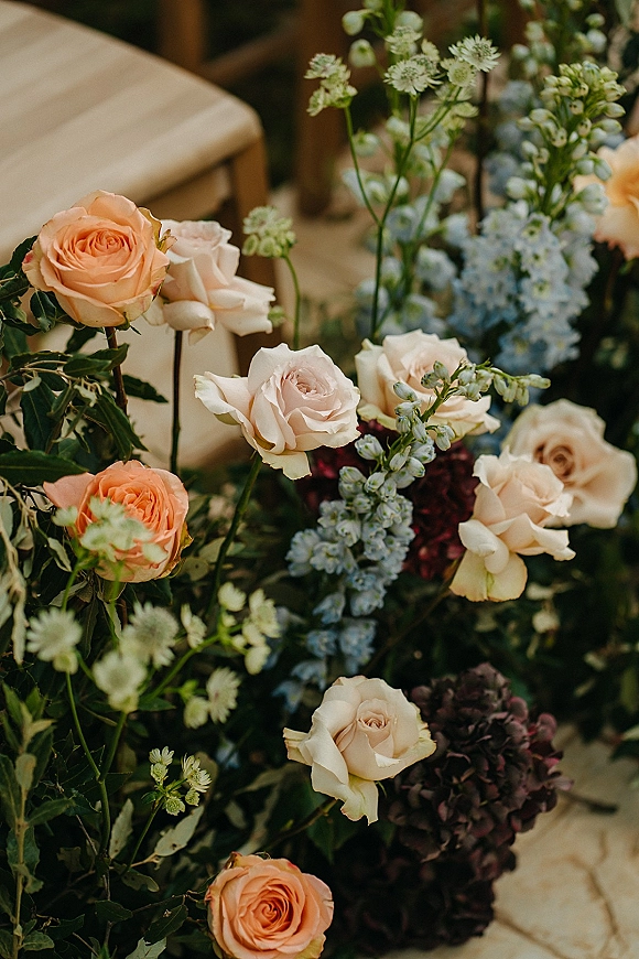 Wedding floral arrangement with peach and cream roses and blue delphinium, nestled by a wooden chair on an indoor floor with greenery