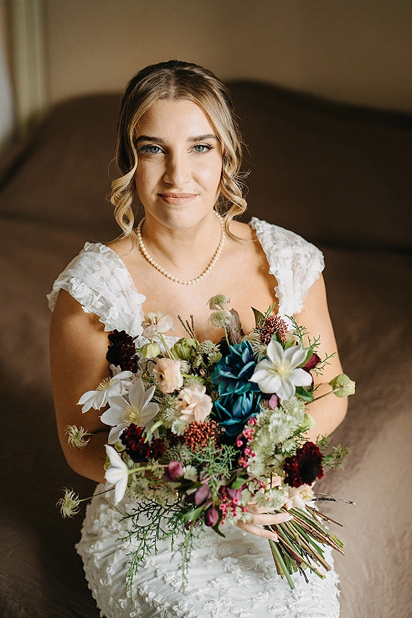 Bridal portrait of a bride holding bouquet in a lace dress with ruffle sleeves and pearl necklace, seated by a sofa indoors