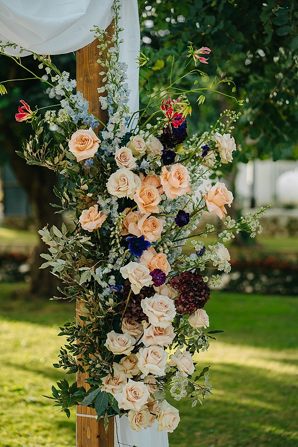 Wedding ceremony arch with wood wedding arch styling, white draping and asymmetrical blush roses, delphinium, and greenery on a garden lawn