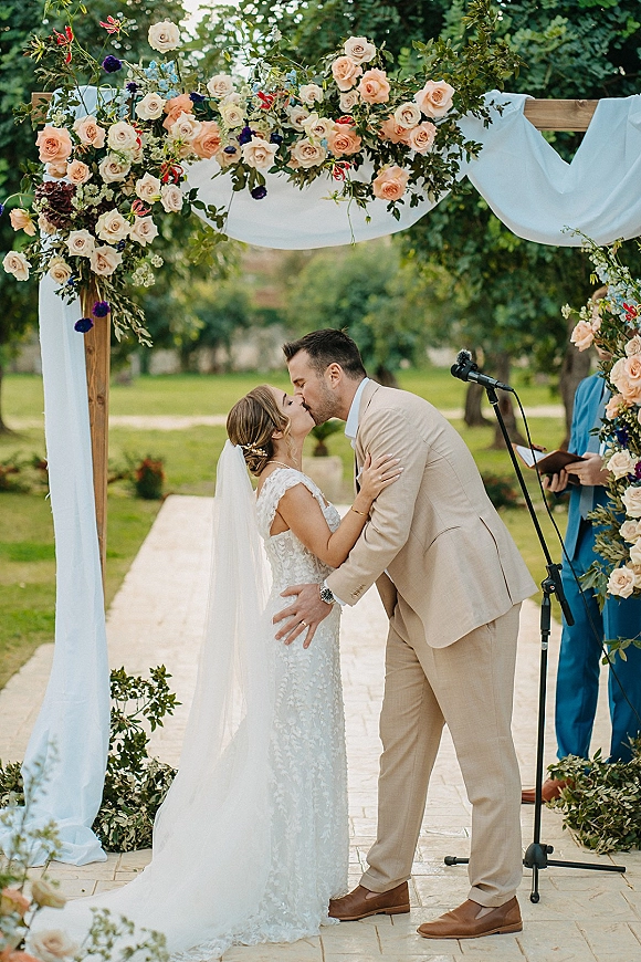 Ceremony kiss as bride and groom embrace under a rose floral arch with white drapery in a garden outdoor wedding ceremony setting