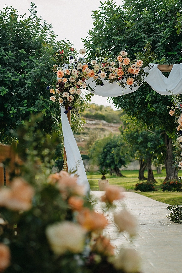 Wedding ceremony arch with a wood outdoor wedding arch draped in white fabric and rose greenery garland in a garden with stone walkway
