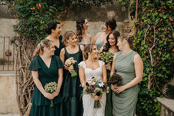 Bridesmaids group photo with bride and bridesmaids laughing, holding bouquets in green dresses before an ivy-covered stone wall outdoors