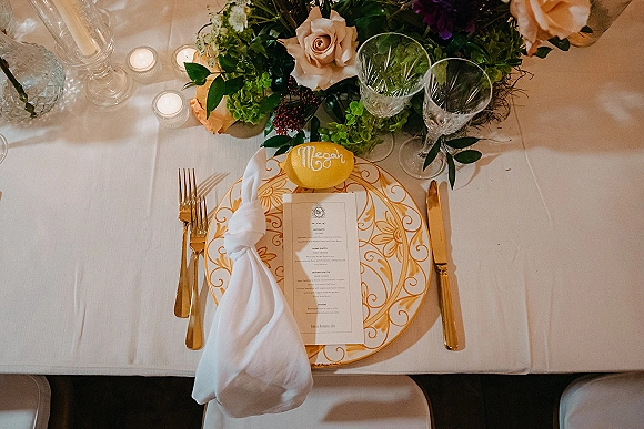 Reception tablescape with wedding place setting on white linen, gold patterned plate, menu card, crystal glasses, roses, votive candles, lemon place card