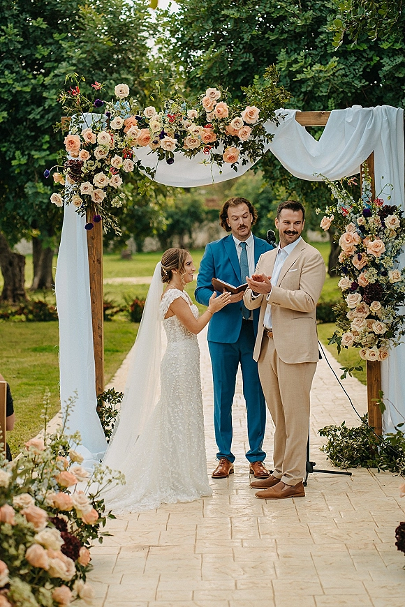 Ceremony moment as bride and groom stand under a floral arch while the officiant speaks beside a microphone on a garden stone aisle