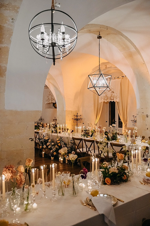 Reception tablescape with candlelit wedding reception glow, taper candle centerpieces, florals and lemons on long tables in a stone-arched hall