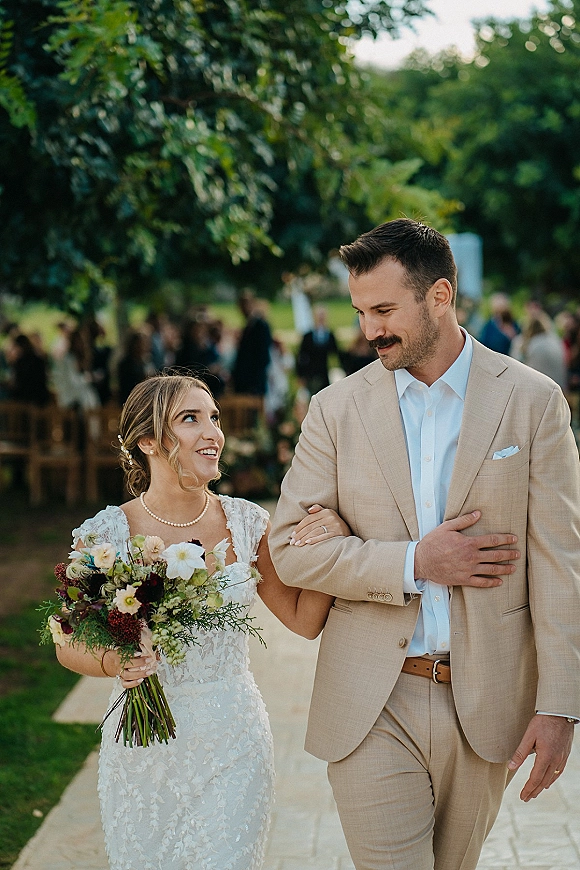 Wedding processional with bride walking down aisle arm in arm with groom, holding wildflower bouquet, guests seated along a garden walkway