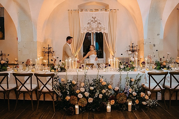 Reception head table with lush floral garland, roses, and taper candles in glass holders, set beneath an arched ceiling with draped curtains.