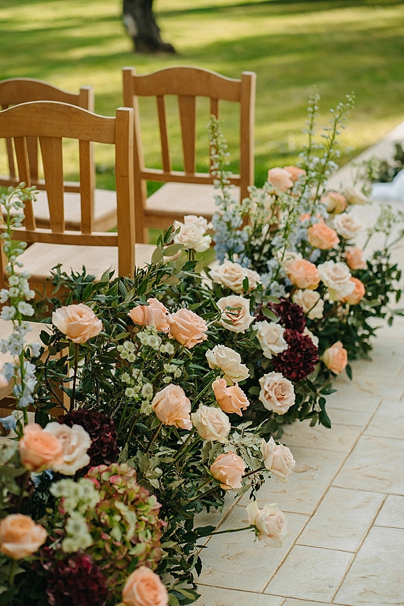 Ceremony aisle flowers form a ground floral aisle with peach and ivory roses, hydrangea, and greenery along a stone walkway by wooden chairs