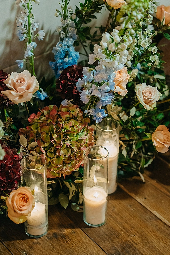 Wedding floral arrangement with ceremony ground florals featuring peach roses, hydrangeas, blue delphinium, and glass hurricane candles on wood floor
