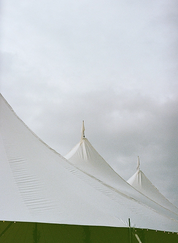Wedding tent with sailcloth peaks and warm string lights, tent poles and pennant flags beneath an overcast sky outdoors