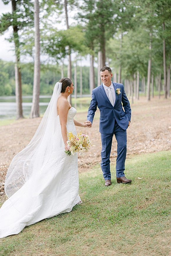 Couple portrait of bride and groom holding hands, her cathedral veil and bouquet flowing as they walk by pine trees near a lakeside lawn