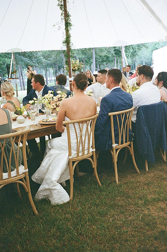 Wedding reception farm table set with wooden chairs, pastel floral centerpiece and candles under string lights in a clear-top tent on grass