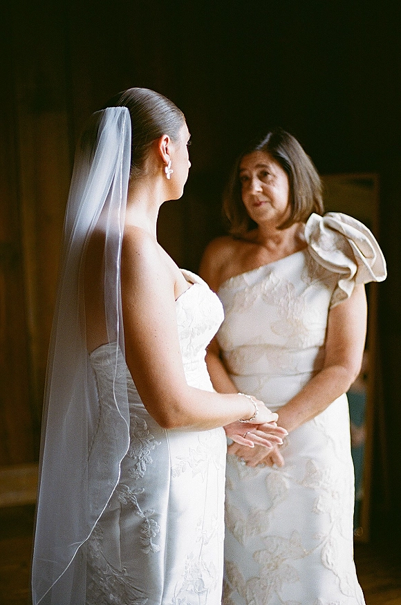 Bride with mother holding hands in a mother daughter wedding moment, veil and lace strapless gown glowing in soft window light indoors
