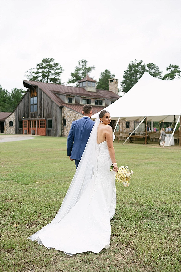 Couple portrait of bride looking back in a strapless lace gown with cathedral veil and bouquet, walking on a lawn near a rustic barn and tent