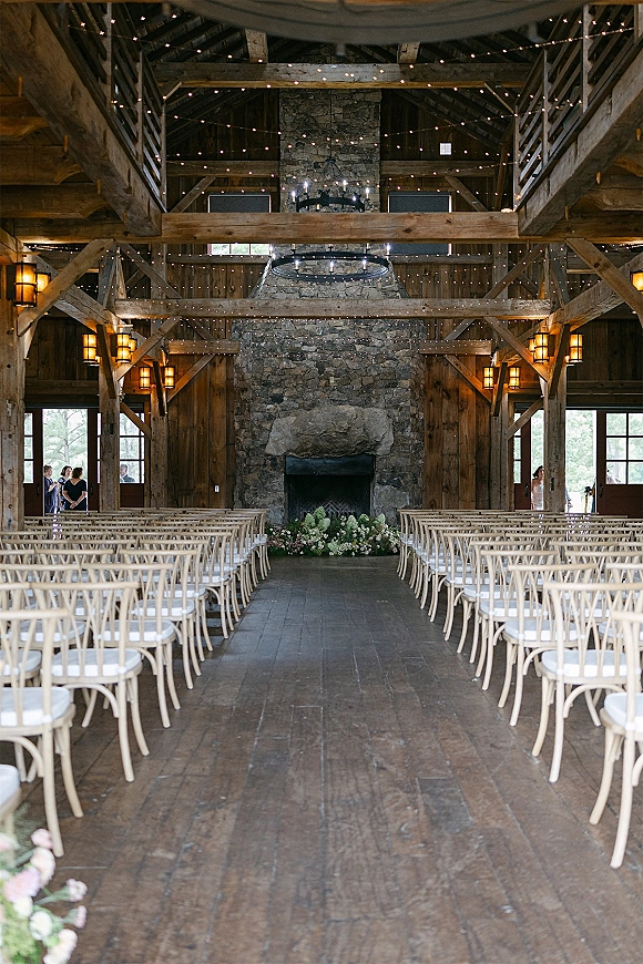 Indoor ceremony setup with barn wedding ceremony chairs lining a wood aisle, string lights and candle chandelier near a stone fireplace altar