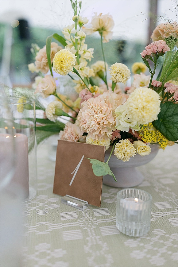 Reception tablescape with a wedding table centerpiece of yellow and blush flowers in a ceramic vase, candlelight on patterned cloth by windows