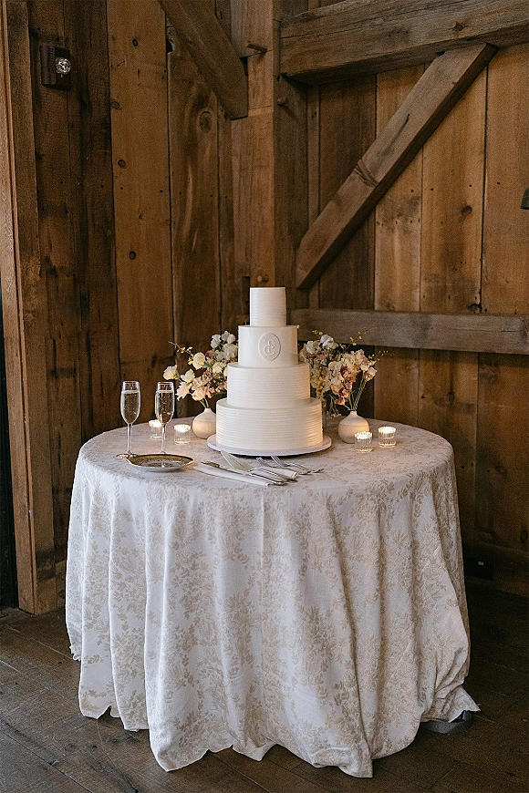 Wedding cake table with a three tier wedding cake on a stand, floral arrangements and candles, champagne flutes, set against a wood barn wall