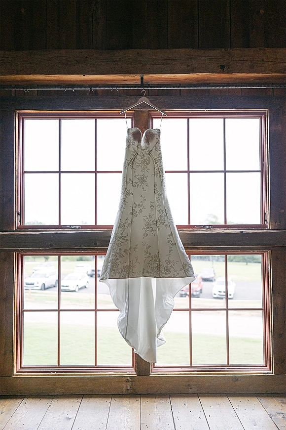 Wedding dress hanging in a barn window, showing a strapless bridal gown with floral lace applique against bright window panes
