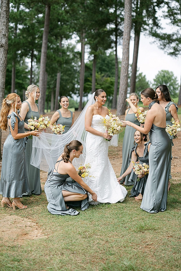 Bridal party portrait of a bride with bridesmaids holding bouquets on a grassy path among pine trees, with a cathedral veil trailing behind