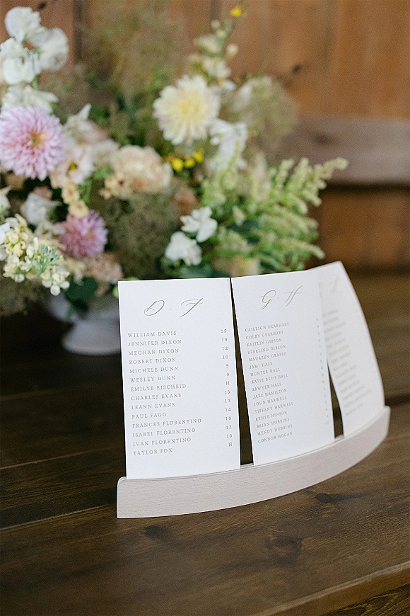 Wedding seating chart with alphabetical escort cards in a wooden holder, calligraphy typography and floral vase on a rustic wood table backdrop