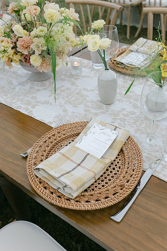 Reception tablescape with wedding place setting featuring rattan charger plates, plaid napkins, menu cards, florals, and candlelight on a wood table outdoors